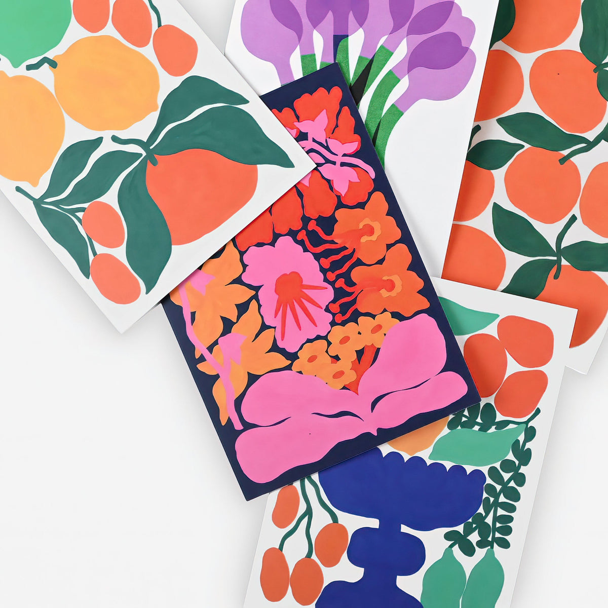 A styled flat-lay of several colorful patterned cards or art prints overlapping on a white background. The designs feature bold abstract floral and fruit motifs in orange, pink, purple, blue, green, and yellow.