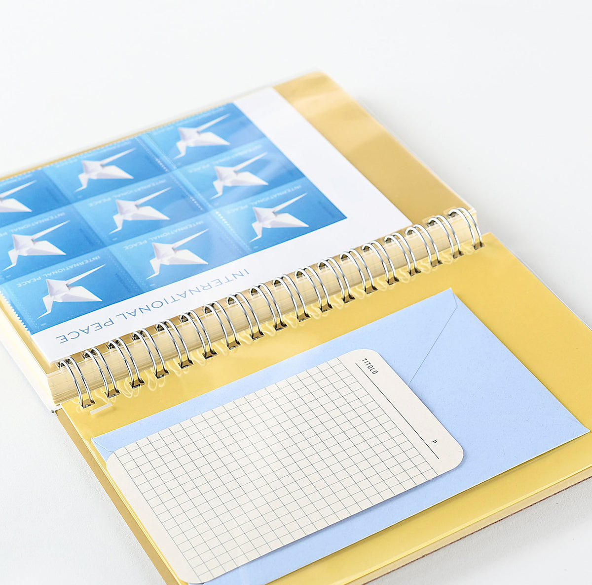 A Rollbahn gold spiral-bound  planner shown open on a white background, featuring a blue 'International Peace' stamp sheet, a light blue paper insert, and a small grid notepad.